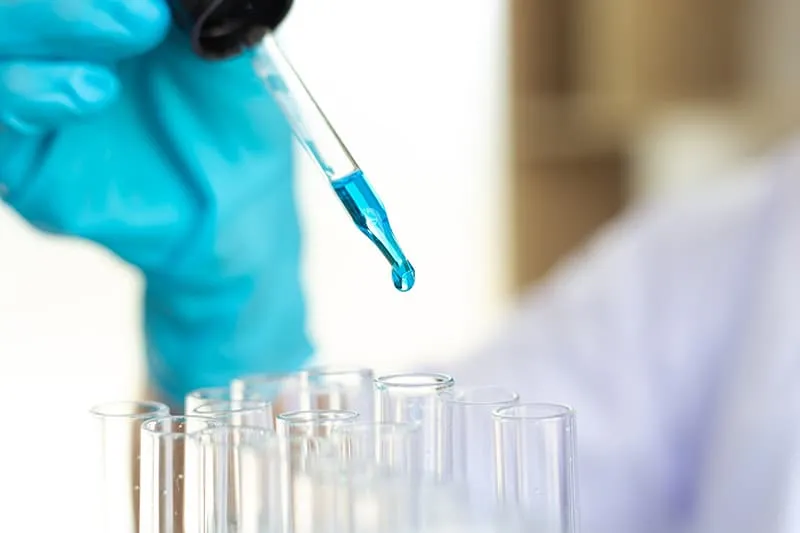A gloved hand uses a glass dropper to release a single drop of blue liquid into one of several clear test tubes in a laboratory setting.