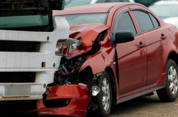 a red sedan has sustained significant front-end damage following a collision with the rear of a white commercial vehicle. The impact has severely crumpled the car's hood and bumper.