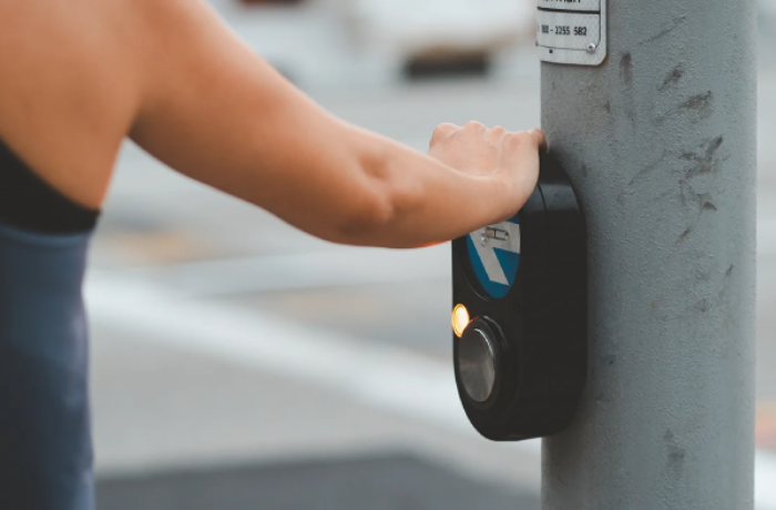 a person's arm is shown reaching out to press a pedestrian crossing button on a metal pole. The background features a blurred street scene with a crosswalk.