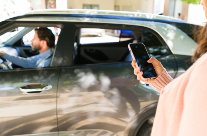 a person in the foreground holds a smartphone while standing near a dark vehicle. Inside the car, a driver is visible through the window, suggesting a rideshare interaction or a coordinated pickup.