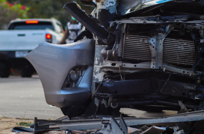 the front end of a vehicle is shown with severe damage, including a mangled bumper and exposed internal components. A white pickup truck is visible in the blurred background.