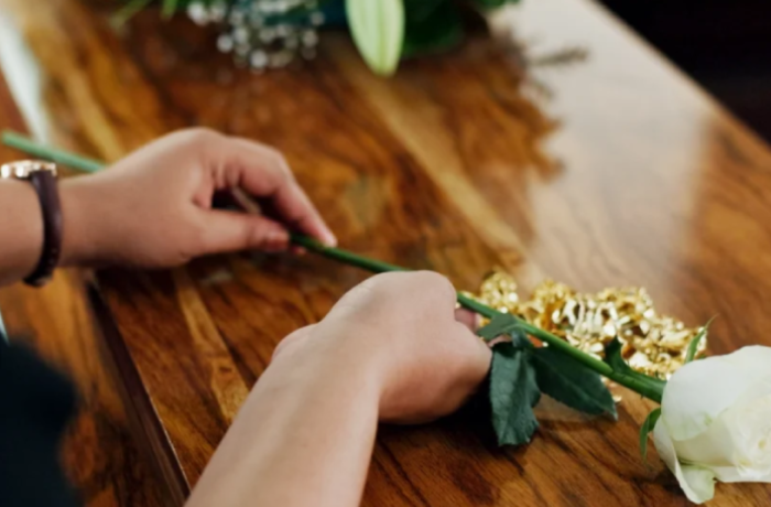 a person in dark clothing rests their hands on a polished wooden casket during a funeral service. A single white rose lies on the lid near their hands, with a larger floral arrangement blurred in the background.