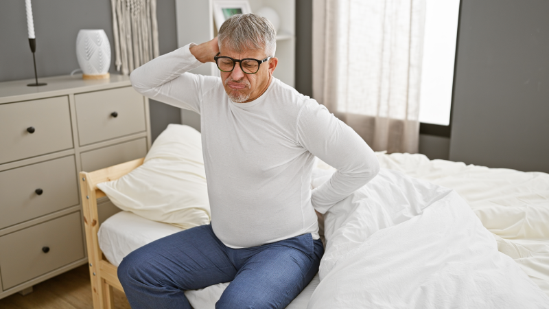 A grey-haired man in pain holding his back while sitting on a bed in a well-lit bedroom