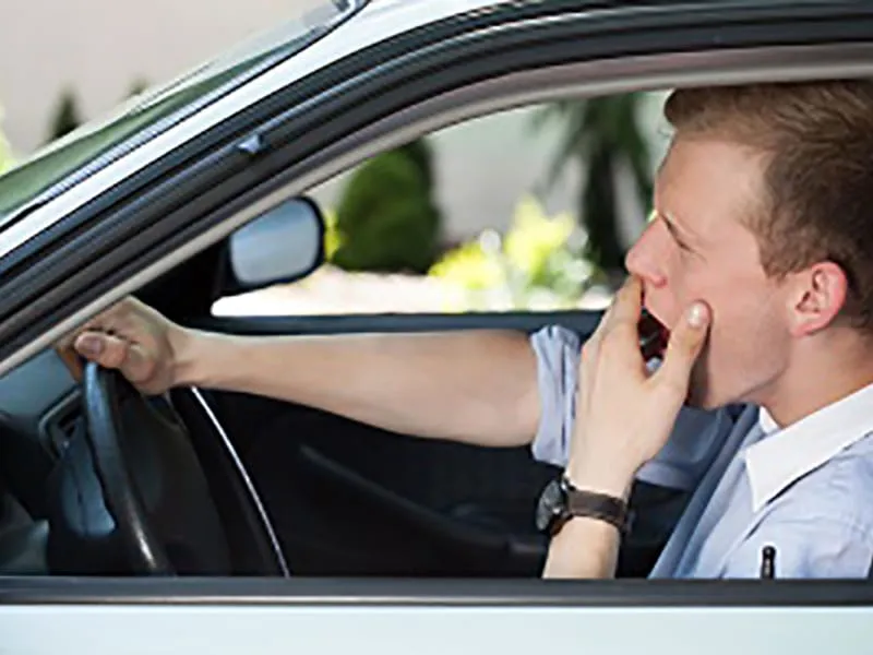 A man sits in the driver's seat of a car, holding the steering wheel with one hand and yawning into his other hand.