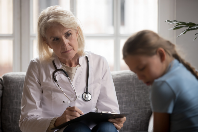 Elderly doctor in white coat sit on couch hold clipboard look at poor small scared closed girl at psychological therapy session meeting, addiction abuse disorders,