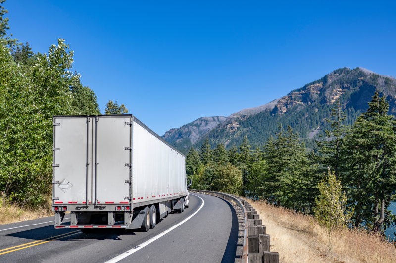 Industrial classic big rig white American semi truck tractor transporting commercial cargo in dry van semi trailer driving on the winding road with green forest