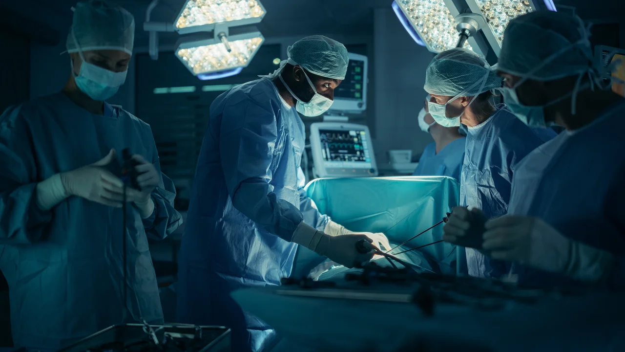a team of surgeons and medical professionals wearing blue scrubs, masks, and caps is shown performing a surgical procedure under bright operating room lights.
